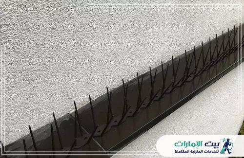 حماية أنيقة ومتينة من الحمام المزعج! تُظهر هذه الصورة تركيب خط طويل من أشواك مكافحة الحمام الداكنة والمثبتة بإحكام على حافة ضيقة بجانب جدار أبيض ذي ملمس. شركة بيت الإمارات 0509783583 توفر حلولاً لا تشوه المظهر الخارجي، بل وتعمل كدرع فعال لردع الطيور. نضمن تركيبًا احترافيًا بجودة عالية.